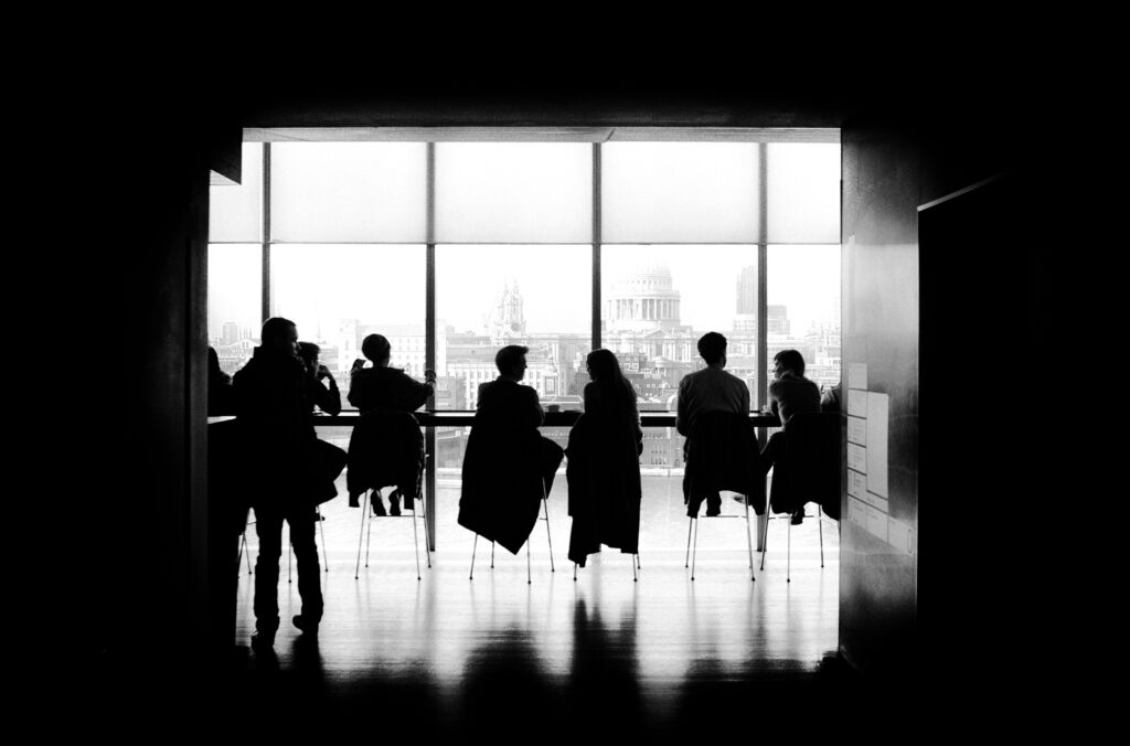 People sitting at a long table in front of a window, facing outside; view of a capitol building