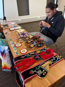 Marcus sits behind a table and fixes a participant's project during a 20 participant workshop. The table in front of him is filled with examples of his handcrafts; a bandolier bag, medicine pouch, scarf tie, headband, earrings, moccasins, ribbon skirt and graduation cap. 
