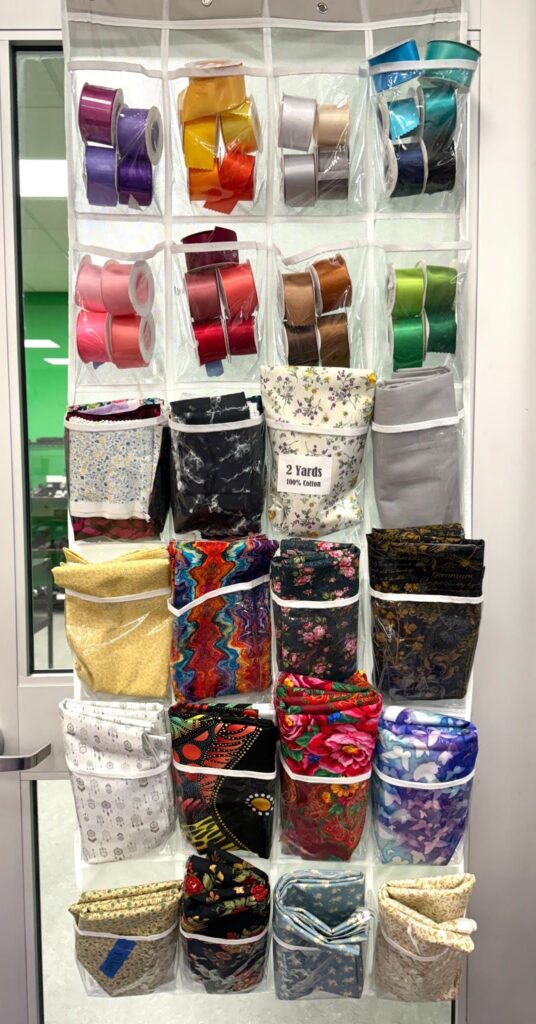A twenty four pocket bag hangs from the back of a door. The first 2 rows of pockets are packed with satin ribbons sorted by color. The next 4 rows down have various 2 yards of cotton fabric in them. These are some of the materials used for sewing ribbon skirts.