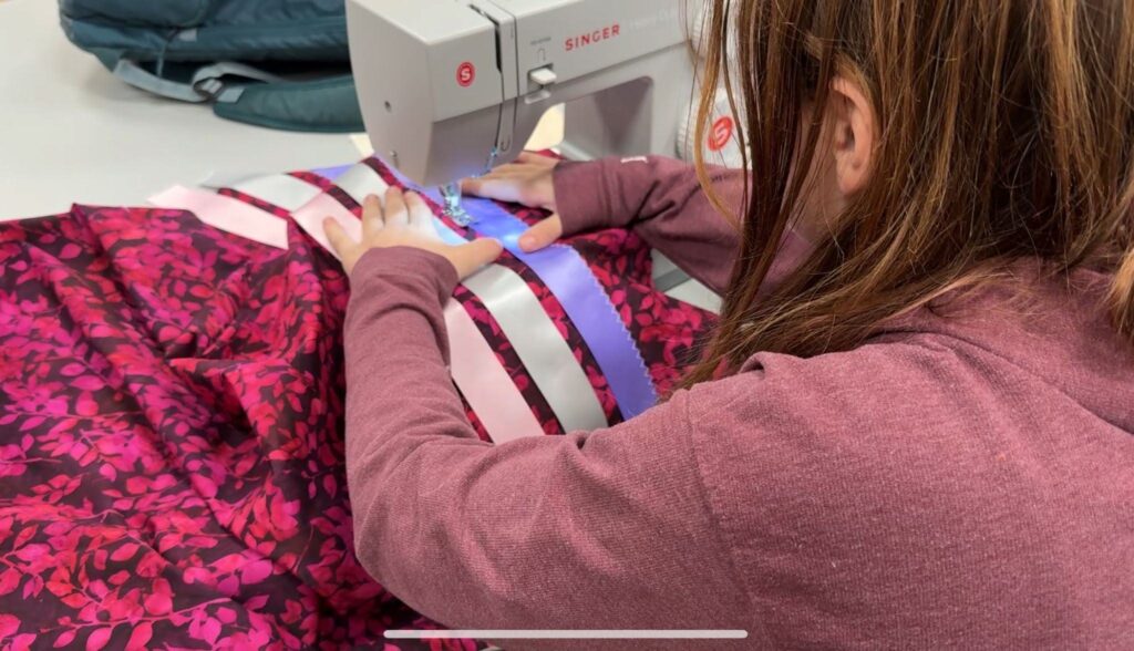 A student is sewing a ribbon skirt using a heavy duty Singer sewing machine. She is sewing down both edges of a purple satin ribbon onto a leaf patterned maroon and hot pink fabric. She will do this with the remaining grey and pink satin ribbons as well. This is her first ribbon skirt.